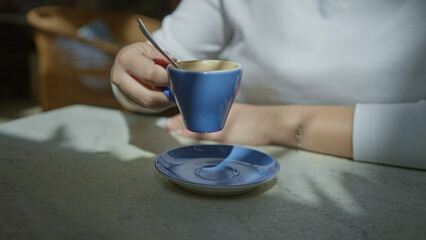 Woman stirring coffee in a blue cup at a mediterranean restaurant, showcasing a relaxed indoor setting bathed in natural light and shadows on a stone table.