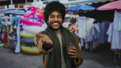 Smiling man in market street holding coffee with vibrant stalls in background reaching playfully forward.