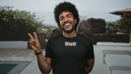 Man with afro smiling in staff uniform outdoors at hotel pool resort showing peace sign surrounded by greenery and white furniture capturing friendly hospitality vibe