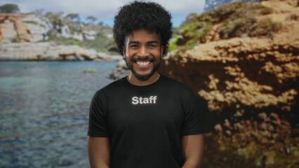 Man smiling in staff uniform at picturesque seaside location with rocky cliffs and clear blue...