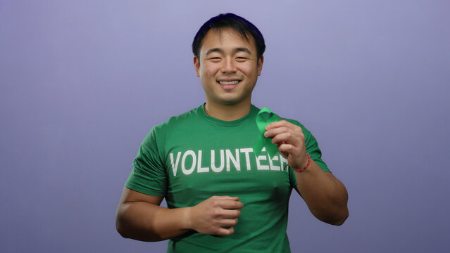 Young man wearing green volunteer shirt holding ribbon against purple background smiling at camera, conveying charity and awareness.
