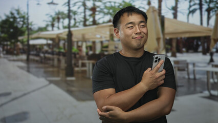 Man holds smartphone against his chest with crossed arms near street cafe seating while glancing...
