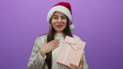 Hispanic woman in santa hat smiling while holding gift against purple background, expressing joy...