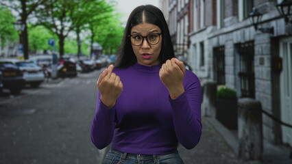 Woman wearing purple sweater pointing fingers to chest on street clad in denim jeans with glasses...