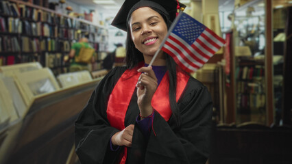 Woman wearing cap and gown smiling while holding american flag in library building; pride...