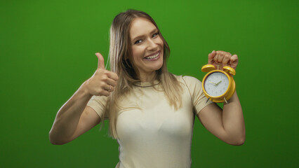 Young woman with blonde hair holding a yellow alarm clock smiling over green isolated background while giving a thumbs up