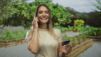 Young woman with blonde hair talking on smartphone and holding a credit card in a sunny park...