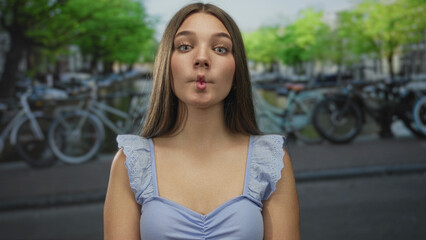 Woman with bare shoulders in pastel blue top puckers lips on street lined with bicycles; playfulness.