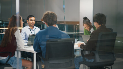 Diverse office teamwork meeting in modern startup workspace with young professionals collaborating around table using laptops and sharing ideas for creative project