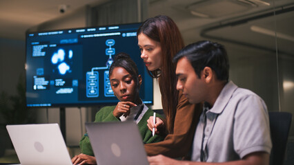 Diverse office teamwork in startup environment as three young professionals collaborate on laptops with focus and determination in modern workspace