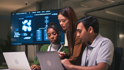 Diverse office teamwork in startup environment as three colleagues collaborate on laptops with focus and determination in modern workspace with digital screen in background