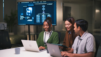 Diverse office teamwork in startup environment as three young professionals collaborate on laptops and digital tablet with flowchart on screen in background