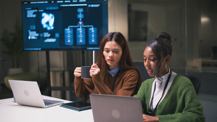 Diverse office teamwork in startup environment with two women collaborating on laptops, discussing data analysis and project strategy in modern workspace with digital screen