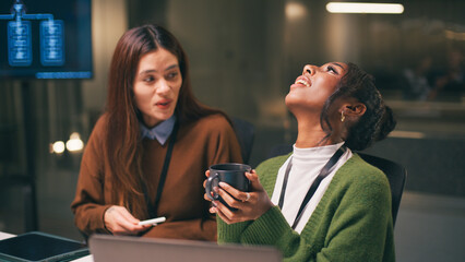 Diverse office teamwork at startup creates joyful atmosphere as two young women share laughter during late work session in modern workspace