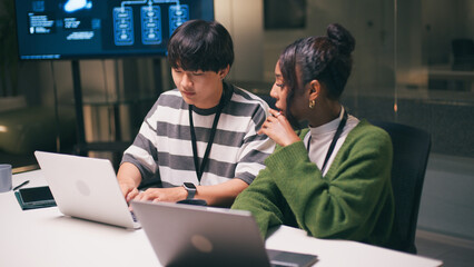 Diverse office teamwork in startup environment as two young professionals collaborate on laptops in...