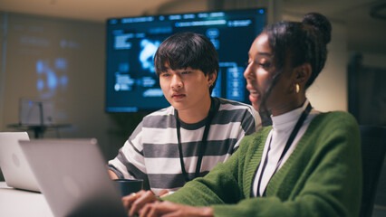 Diverse office environment with teamwork as two startup colleagues collaborate on laptops in modern...
