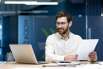 Smiling young businessman holding an online meeting, sitting at a table in the office, working with documents and looking at a laptop
