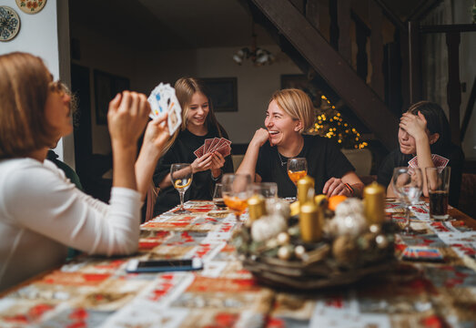 Cheerfully warm home togetherness cozy scene of family playing cards together in home kitchen. Girls and boys smiling and laughing with moms. Holidays time spending and relations concept image.