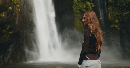 Fototapeta premium A woman stands in front of a majestic Bali waterfall, surrounded by lush greenery. The water cascades down, creating a breathtaking natural spectacle.