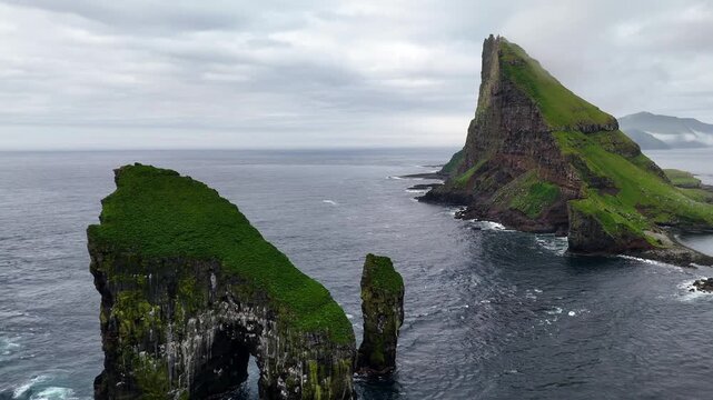Cinematic aerial view of Drangarnir sea stacks rising dramatically from the Atlantic Ocean near V&aacute;gar, Faroe Islands, showcasing rugged cliffs, lush green slopes, and misty Nordic seascape