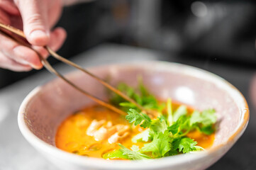 Close-up of hand garnishing spicy vegetable soup with fresh cilantro using chopsticks. Vibrant orange broth, mushrooms, and herbs in a restaurant or culinary setting