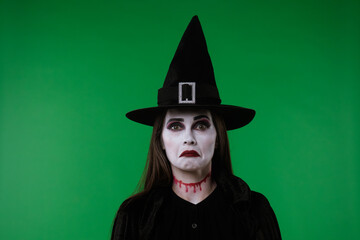 A witch with vampire makeup, wearing a black cloak and hat, looks at the camera and appears very...