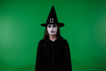 A witch with vampire makeup, wearing a black cloak and hat, looks at the camera and appears very...