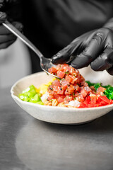 Chef wearing black gloves prepares a poke bowl with diced raw tuna, bell peppers, and rice on a stainless steel surface in a professional kitchen