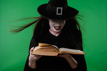 A vampire witch in a black cloak casts spells over a magic book against the green background of a...