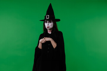 A witch with vampire makeup, wearing a black cloak and hat, looks at the camera and appears very...