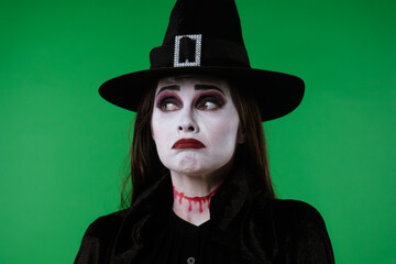A witch with vampire makeup, wearing a black cloak and hat, looks at the camera and appears very...