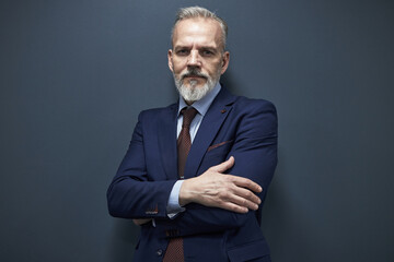 Portrait of mature Caucasian man with gray beard and mustache standing with arms crossed looking into camera wearing formal business suit against plain background