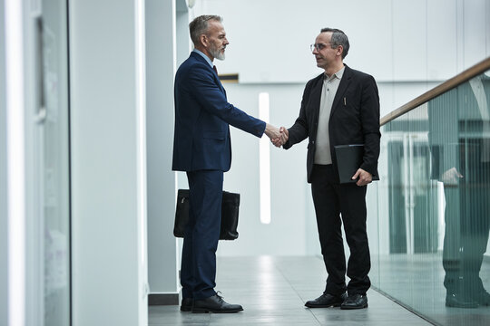 Two middle aged Caucasian men shaking hands in modern office hallway, both standing and making eye contact, one holding briefcase, other holding tablet, business agreement concept