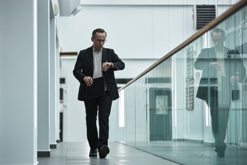Middle aged Caucasian businessman walking in modern office hallway, checking wristwatch, holding...
