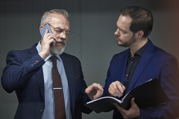 Middle aged Caucasian male executive talking on smartphone while standing next to young partner...