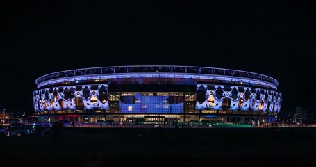 Cycling LED shapes covering stadium facade at night, with glass entrance glow, vehicle light trails - Powered by Adobe