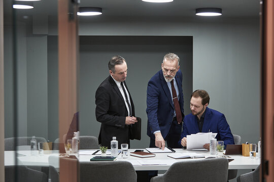Three Caucasian men, two mature nd one young, collaborating in modern conference room, discussing documents and analyzing financial data in paperwork during business meeting