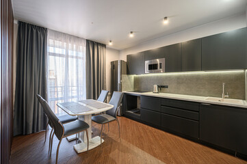 Modern kitchen with dark cabinets, a white dining table with four chairs, and hardwood floors. Natural light streams through sheer curtains