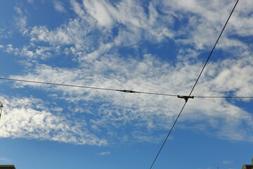 Blue sky with white clouds intersected by tram or trolleybus wires, with city buildings below. Urban infrastructure and abstract cityscape concept.
