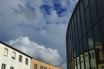 Modern glass building reflecting traditional residential houses under dramatic cloudy sky. Urban architecture contrast and cityscape design concept.