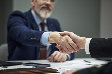 Mature businessman with beard shaking hands with young male partner across desk in office during...