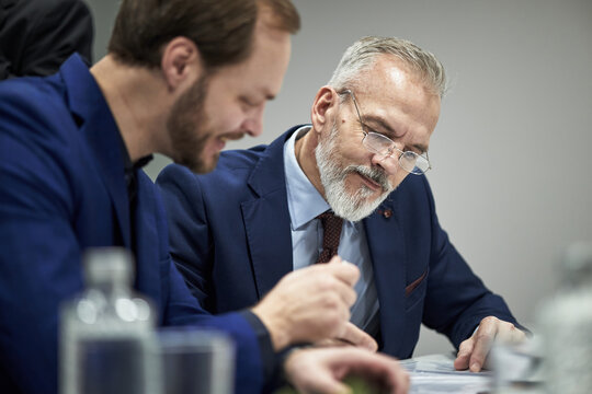 Middle aged Caucasian man with gray beard sitting beside young male partner, both wearing suits, reviewing documents together at table in office, conducting negotiations on contract terms