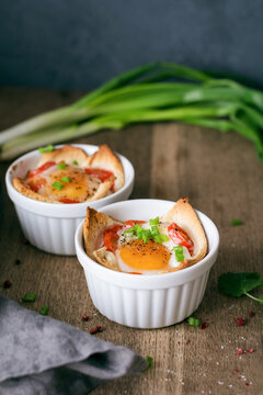 Close-up of two Baked eggs, tomatoes, spring onions and bread ramekins on a wooden table