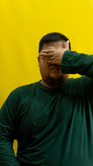 Man in green long sleeve shirt covering eyes with hand, isolated on yellow background, expressive...