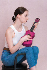 Serious young female musician playing ukulele, sitting on bar stool and looking away while plucking...