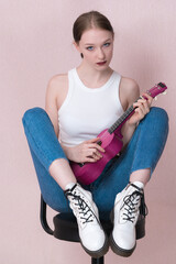 Young female musician plays ukulele, sits on bar stool and looks straight into camera with blank expression. Beautiful woman is wearing casual clothing white tank top, blue skinny jeans and boots