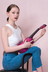 Woman holding ukulele tunes strings of her musical instrument and looks at camera with blank expression. Musician sitting on bar stool. Woman in casual clothing - white tank top and tight jeans