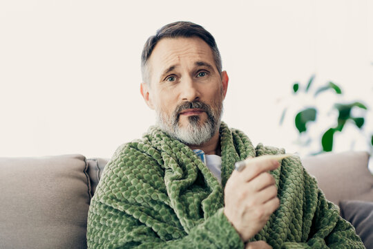 Senior man wearing casual attire while sitting on a sofa indoors, holding a thermometer, and appearing reflective