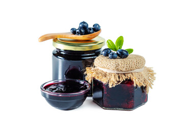 Jar and bowl of blueberry jam with berries, leaves and wooden spoon isolated on white background.