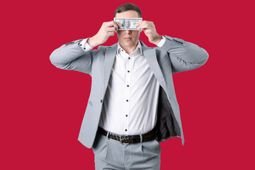 A man in a grey suit, white shirt and brown belt holds a banknote in front of his eyes against a bright red background.